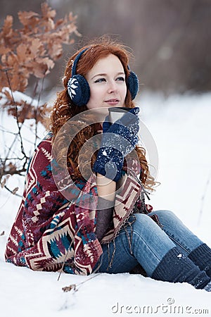 Dreamly young red-haired woman drinking a hot drink from a mug in the winter park. Stock Photo