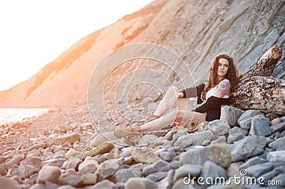 Dreamy lonely brunette hippie in the style of bokho chic sits on gray stones at the sea shore in the rays of the setting sun Stock Photo