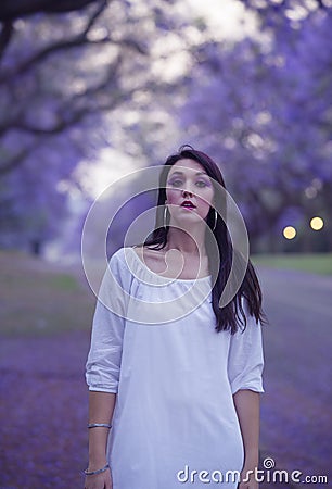 Dreamy image of beautiful woman in white dress walking in street surrounded by purple Jacaranda trees Stock Photo
