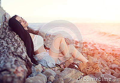 Dreamy lonely brunette hippie in the style of bokho chic sits on gray stones at the sea shore in the rays of the setting sun Stock Photo