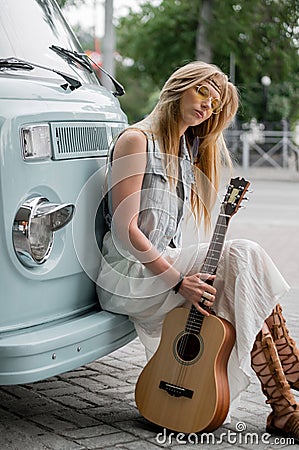 Dreamy woman sits by the camper. A hippie girl holding a guitar and leaning on a classic travel van. Stock Photo