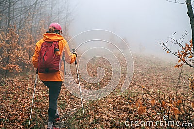 Dressed bright orange jacket young female backpacker walking by the touristic path using trekking poles in autumn foggy forest. Stock Photo