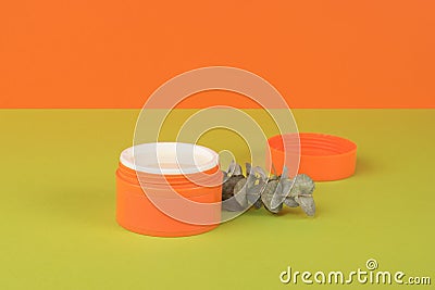 A dried sprig of eucalyptus near an open jar with cosmetic cream on an orange and green background Stock Photo