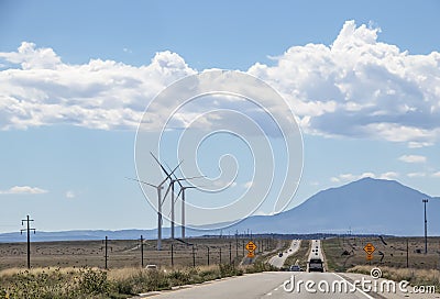 Driving on a long straight road with heat shimmer toward mountains - wind turbines on one side and signs that say Gusty Winds Area Stock Photo