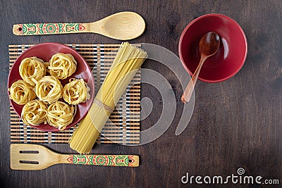Dry fettuccine pasta on a red ceramic plate on a softly blurred background of patterned kitchen utensils and bamboo napkins and Stock Photo