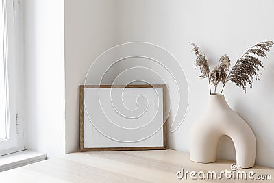 Dry grass, reed plant bouquet in modern ceramic vase. Empty photo frame mockup against white wall on wooden table near Stock Photo