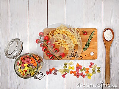 Dry Italian pasta fettuccini and farfalle with tomatoes, rosemary, mixture of peppers, glass jar, spoon with flour and board Stock Photo