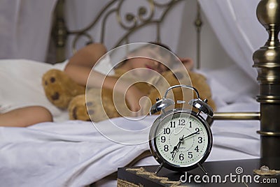 Early awakening. Alarm clock standing on bedside table. Wake up of an asleep young girl holding teddy bear in bed on a Stock Photo