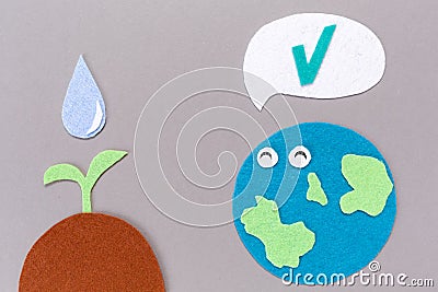 Earth Day. Cutted out of felt the planet Earth with smiling emoticon and soil with a watered plant sprout. Gray background. Flat Stock Photo