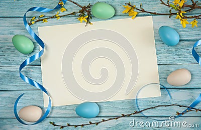 A blank sheet of paper, branches of forsythia with yellow flowers, ribbons and Easter eggs on a light blue wooden background. Stock Photo