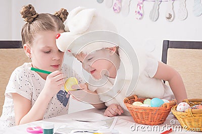 Easter festival: children paint easter eggs at the table. A boy in a rabbit costume looks cunningly at the work of the older siste Stock Photo