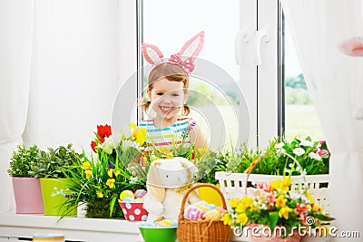 Easter. happy child girl with bunny ears and colorful eggs sitting at window in flowers Stock Photo