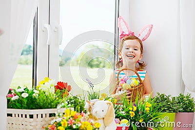 Easter. happy child girl with bunny ears and colorful eggs sitting at window in flowers Stock Photo