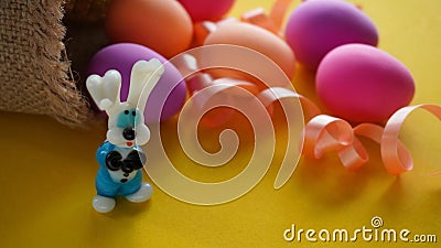 Easter with a hare glass figurine, colored chicken eggs and a pink ribbon on a yellow background Stock Photo