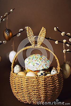 Easter holidays, cute basket with bunny ears on brown background. Easter cakes with colored sprinkles, colored golden eggs, and Stock Photo
