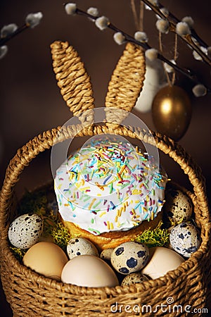 Easter holidays, cute basket with bunny ears on brown background. Easter cakes with colored sprinkles, colored golden eggs, and Stock Photo