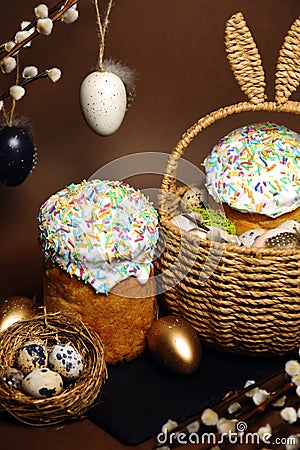 Easter holidays, cute basket with bunny ears on brown background. Easter cakes with colored sprinkles, colored golden eggs, and Stock Photo