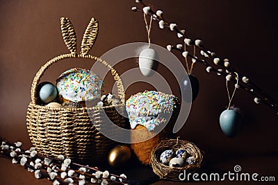 Easter holidays, cute basket with bunny ears on brown background. Easter cakes with colored sprinkles, colored golden eggs, and Stock Photo