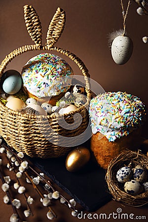 Easter holidays, cute basket with bunny ears on brown background. Easter cakes with colored sprinkles, colored golden eggs, and Stock Photo