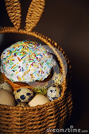 Easter holidays, cute basket with bunny ears on brown background. Easter cakes with colored sprinkles, colored golden eggs, and Stock Photo