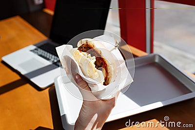 Eating at work place - fast food. burger near laptop. lunch break while you work or sharing online. the closeup hand Stock Photo