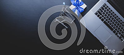 Ecommerce. Laptop computer, shopping trolley and white gift with blue ribbon on dark background. Website retail business Stock Photo