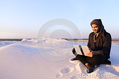 Educated Arab student uses laptop and works sitting on sand amid Stock Photo