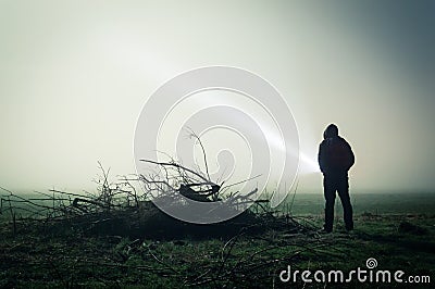 An eerie silhouette of a lone hooded figure in a field on a foggy night with a torch. With a dark edit Stock Photo