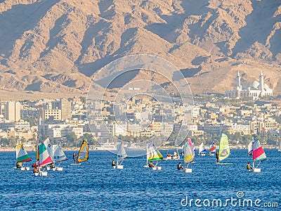 People move in boats with a sail and windsurfing in the Red Sea. Background - the mountains and the city of Aqaba Editorial Stock Photo