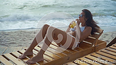 Eine Schönheit in einem weißen Badeanzug und in den Sonnenbrillen liegt auf einem hölzernen Klappstuhl auf dem Seestrand und trin stock video Eine Schönheit in einem weißen Badeanzug und in den Sonnenbrillen liegt auf einem hölzernen Klappstuhl auf dem Seestrand und trin stock video