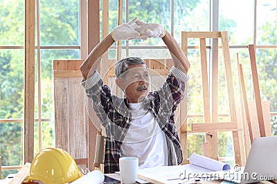 Elderly Asian carpenter with mustache taking a break and drinking a coffee, relaxing senior craftsman setting in working desk at Stock Photo