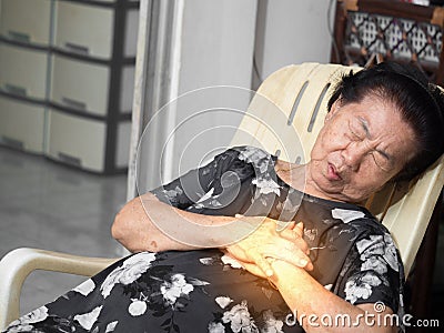 Elderly asian woman sitting on a chair at living room with heart attacks.Both woman`s hands on breast because of hard breathing Stock Photo