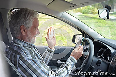 Elderly Caucasian man stops driving the car to answer the video call on the phone. Smiling bearded elderly grandfather holding Stock Photo