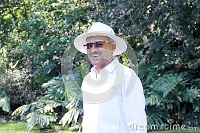 Elderly dark-skinned Latino man in his 60s relaxed in his retirement dressed in a guayabera, hat and sunglasses walks in the park Stock Photo
