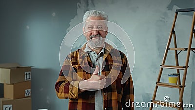 Elderly gray haired man with beard is looking at camera, showing thumbs up and smiling. Portrait of pensioner in plaid Stock Photo