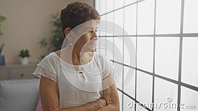 Elderly hispanic woman with short hair stands with her arms crossed in a living room with potted plants and large windows Stock Photo