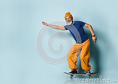 Elderly male in t-shirt, orange pants, hat, gumshoes. Riding black skateboard posing sideways on blue background. Full length Stock Photo