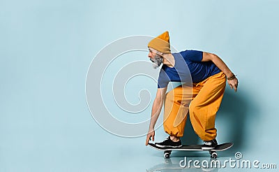Elderly man in t-shirt, orange pants, hat, gumshoes. Riding black skateboard, posing sideways on blue background. Full length Stock Photo