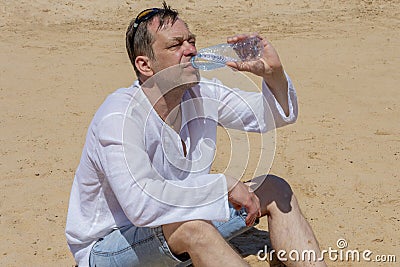 An elderly man 45-50 years old greedily drinks water from a plastic bottle sitting on the sand in the desert. Concept: thirst and Stock Photo