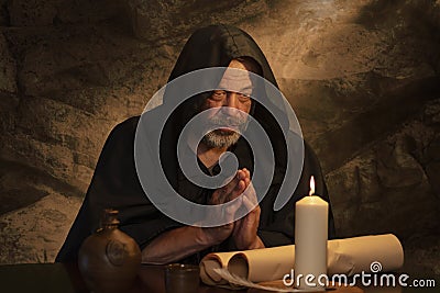 An elderly monk in a black cassock praying, hands folded in prayer, a dark cell, a candle burning on a table. Stock Photo