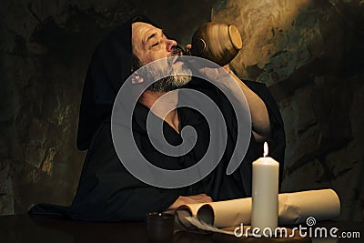 An elderly monk drinking from a clay jug, a dark and smoky stone cell, a candle burning. Stock Photo