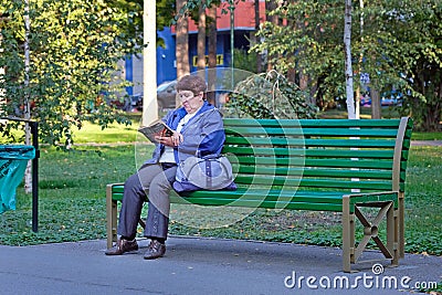 An elderly woman in a blue jacket is reading a book on a bench. Editorial Stock Photo