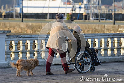 Elderly woman and a disabled man in a wheelchair walk along the street rear view Editorial Stock Photo