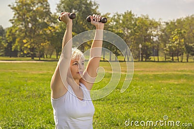 Elderly woman lifts dumbbells while doing fitness in a city park against the backdrop of mountains Stock Photo