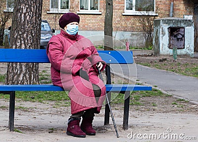 An elderly woman in a medical mask sits on a bench alone Editorial Stock Photo
