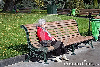 An elderly woman in a red jacket sits on a bench in a park. Editorial Stock Photo