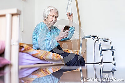 Elderly 96 years old woman reading phone message while sitting on medical bed supporting her by holder. Stock Photo