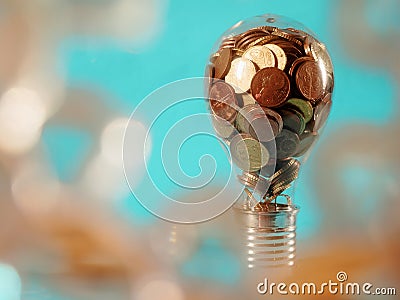 Electric bulb full of coins on blue background in dreamy bokeh foreground. Growing cost of light and energy concept. Payment for Stock Photo