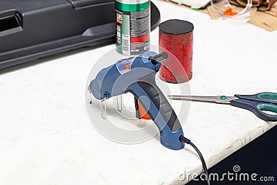 Electric glue gun for squeezing blue glue on the workbench for needlework in the workshop next to scissors and weighting agent for Stock Photo