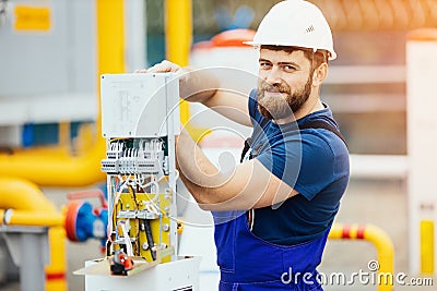 Electrician in a helmet and overalls works adjusts the equipment looks into the camera and smiles. Stock Photo